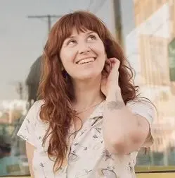 A woman with red hair is smiling and holding her hand to the side of her head.